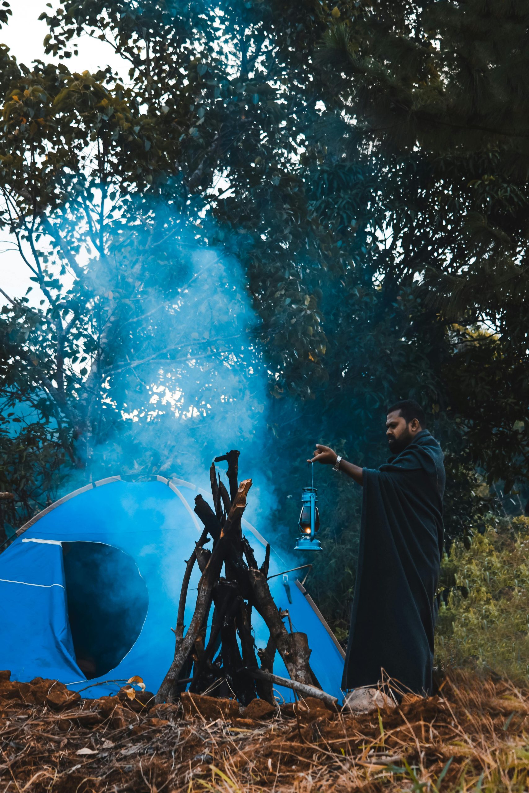 A man holds a lantern near a campfire beside a tent in a forest setting.